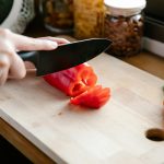 Crop anonymous person slicing ripe fresh red capsicum on chopping board on kitchen counter